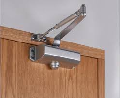 A heavy-duty hydraulic door closer mounted to the top of a commercial entry door and its frame. The silver metal cylinder contains oil and a spring mechanism connected to an adjustable arm, ensuring the door closes smoothly and automatically after being opened.